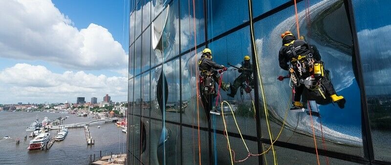 Trzech alpinistów przemysłowych myje okna na wysokościowcu z widokiem na miasto i port, odbicie chmur w szklanej fasadzie.