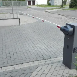 Szara automatyczna bariera wjazdowa z czerwoną naklejką na ramieniu, obok kostka brukowa, krzew i fragment ogrodzenia panelowego w tle.