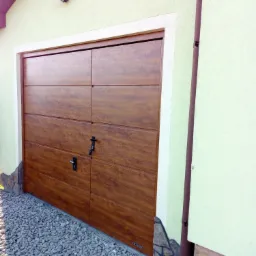 Nowoczesna, segmentowa brama garażowa w odcieniach brązu, z widocznymi panelami i czarnymi uchwytami, zamontowana w jasnym budynku, z kamiennym wykończeniem u podstawy i oświetleniem zewnętrznym.