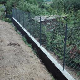 ALFABRAM SP. Z O.O. - Ukończone ogrodzenie z siatki na podmurówce betonowej, widok z dołu zbocza, w tle zieleń drzew i zabudowania.