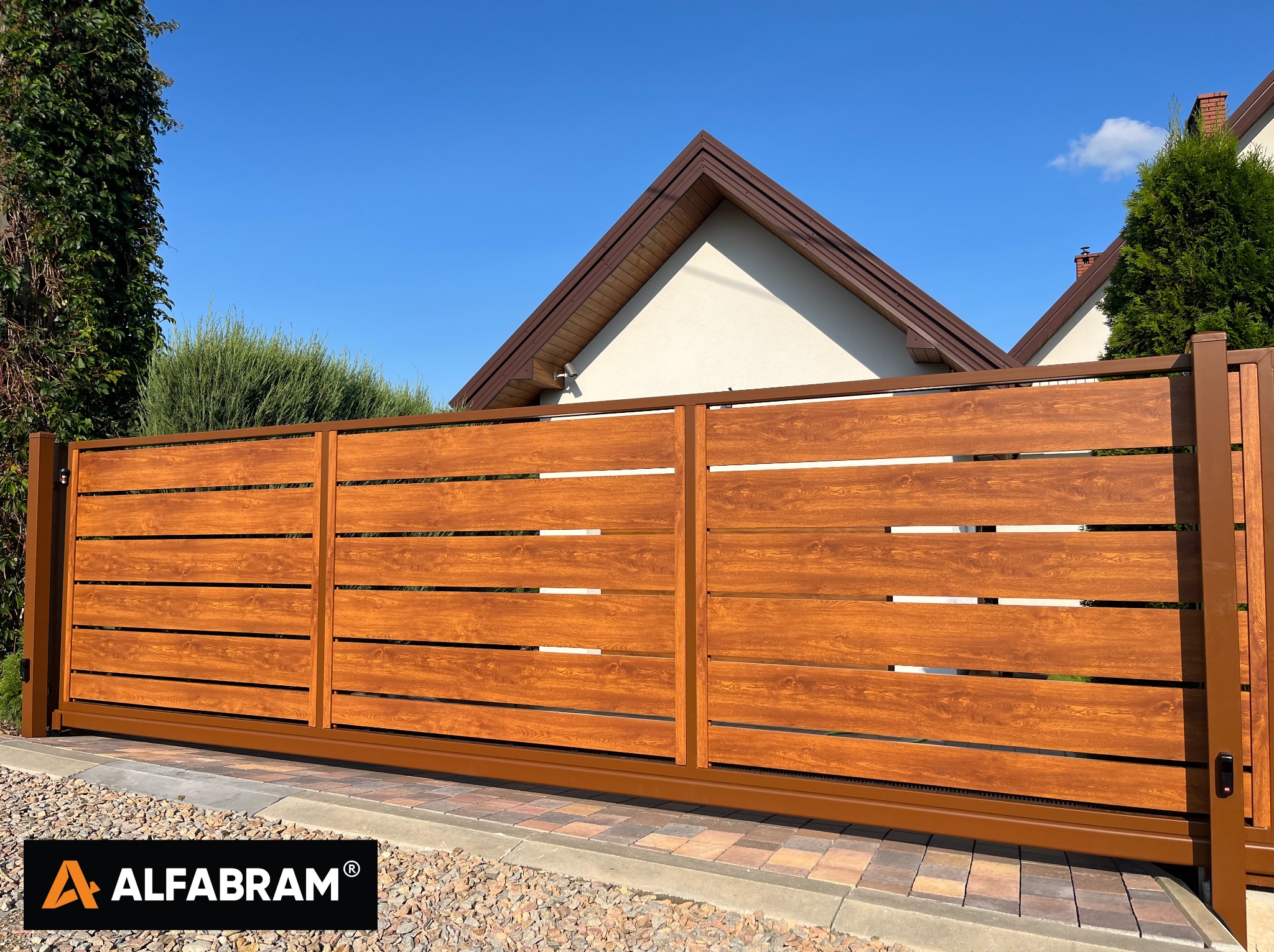 Nowoczesna brama przesuwna z poziomych paneli drewnopodobnych w kolorze orzechowym, z automatyką, zamontowana w Nowym Sączu, na tle domu z brązowym dachem i zieleni.