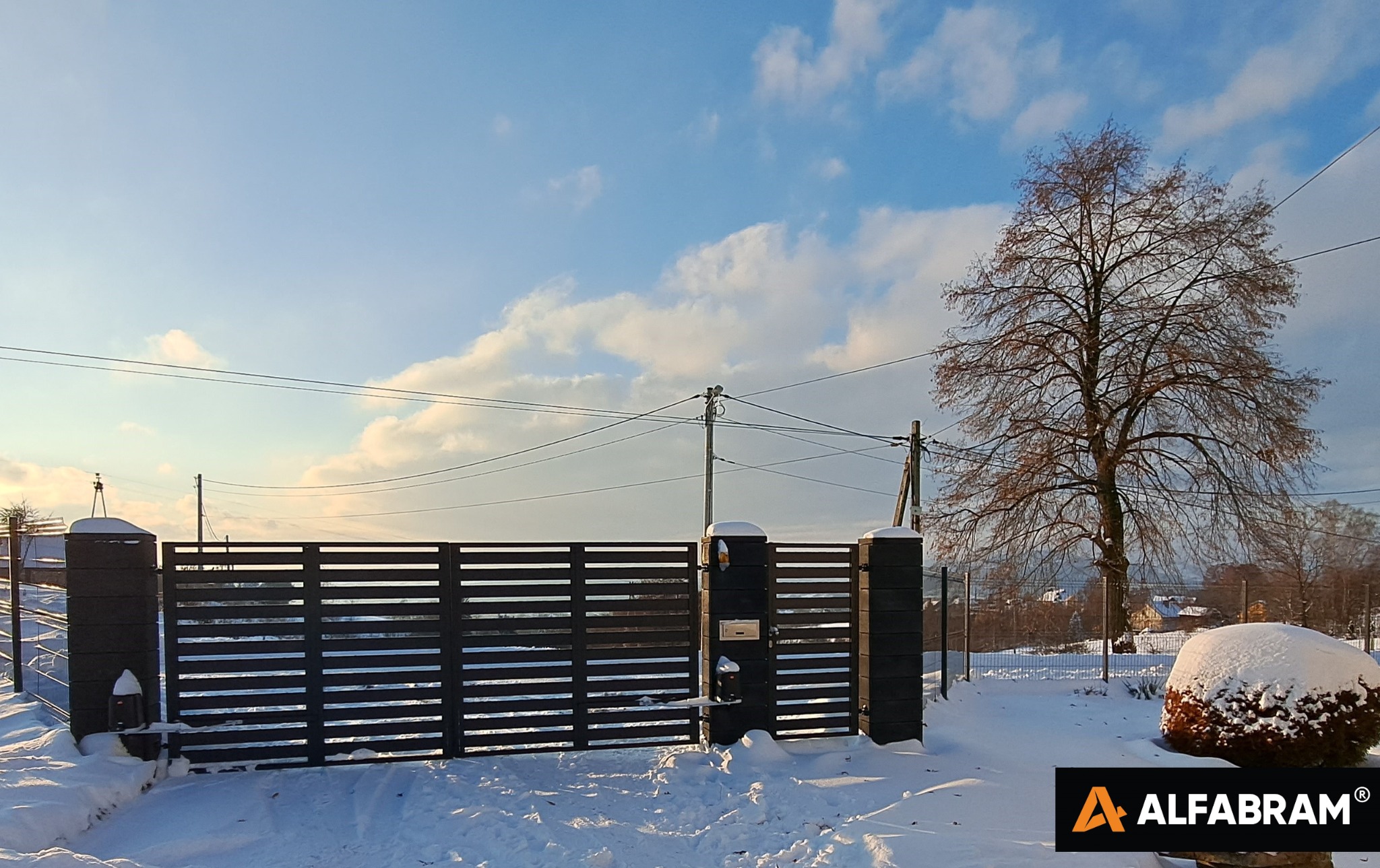 Zimowy krajobraz z nowoczesną, poziomą bramą wjazdową z ciemnego drewna, osadzoną na masywnych, betonowych słupkach, pokrytych śniegiem, z skrzynką pocztową wbudowaną w jeden ze słupków; w tle...