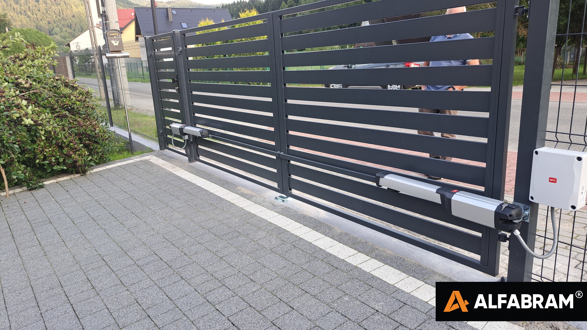 Nowoczesna, pozioma, panelowa brama wjazdowa w kolorze grafitowym z automatyką BFT, widoczna kostka brukowa i krzew z owocami w tle.