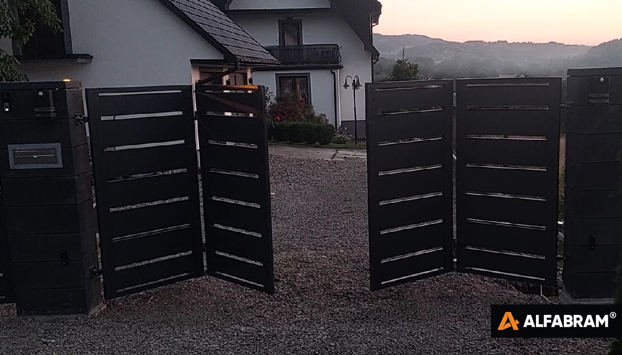 Nowoczesna, otwarta brama wjazdowa z poziomymi przerwami, osadzona na słupkach z szarego kamienia z zamontowaną skrzynką na listy, widok na dom i zalesione wzgórza w tle, logo producenta w prawym...