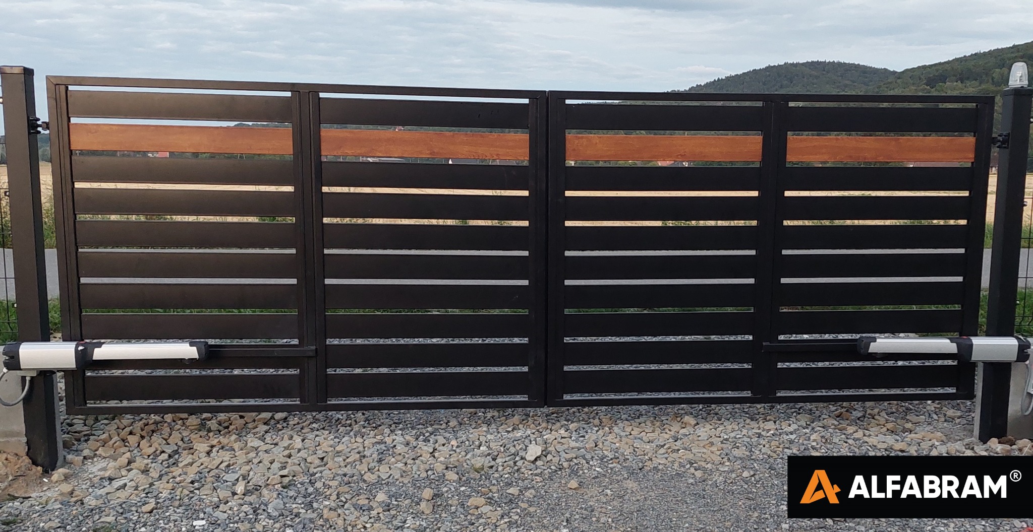 Nowoczesna, dwuskrzydłowa brama wjazdowa z czarnymi, poziomymi panelami i drewnianym akcentem, automatyka zamontowana na słupkach, żwirowe podłoże, w tle wzgórza.