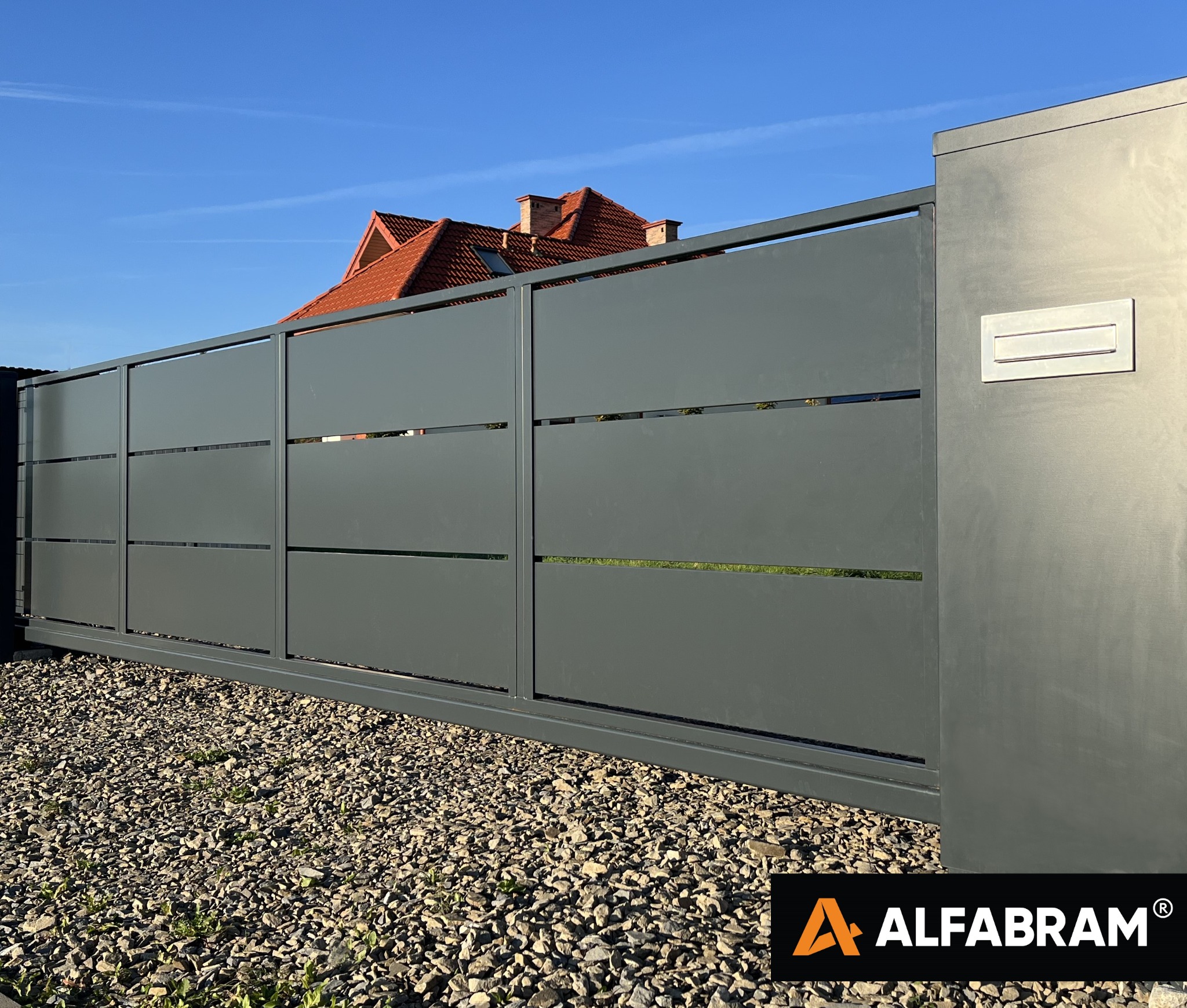 Nowoczesna, przesuwna brama wjazdowa z poziomymi szczelinami, szara, z wbudowaną skrzynką na listy, na tle żwirowego podjazdu i domu z czerwoną dachówką.