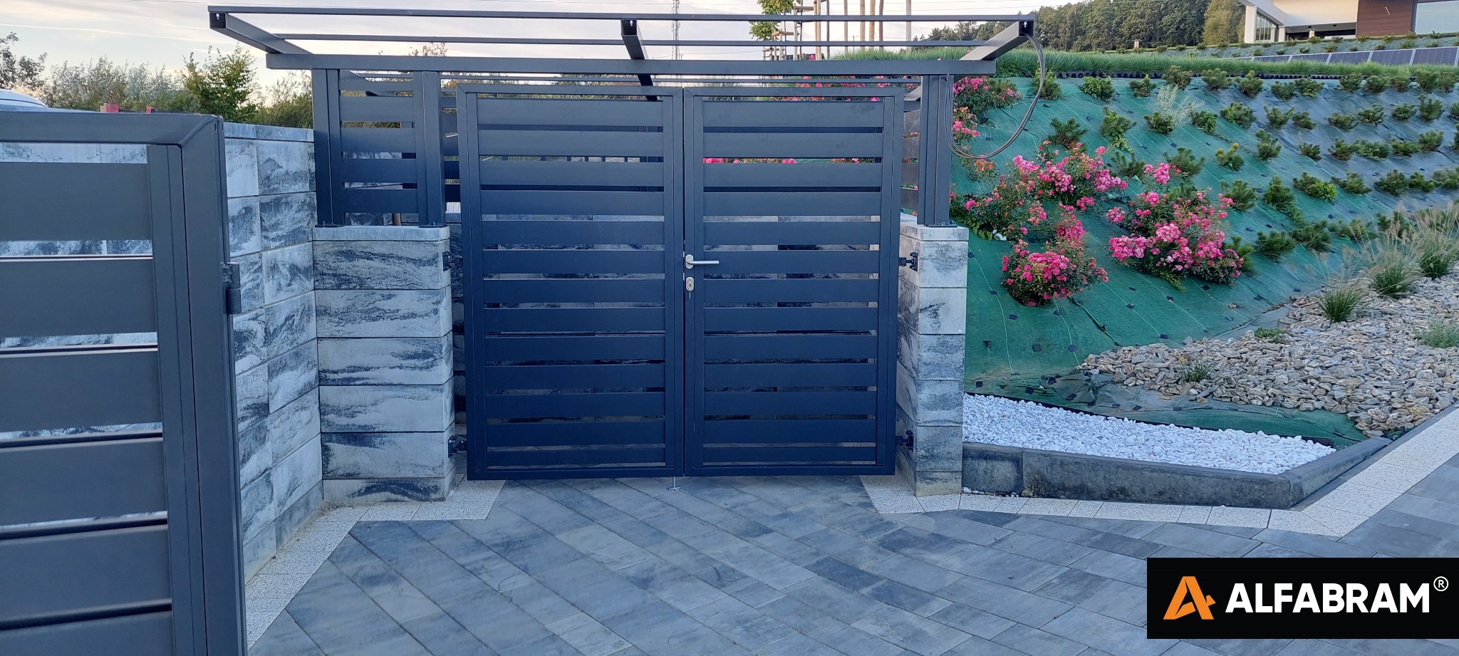 Nowoczesna, pozioma brama wjazdowa z szarych paneli, osadzona na kamiennych słupkach, z widoczną zadaszoną konstrukcją. W tle zielony nasyp z różowymi kwiatami i białymi kamieniami.