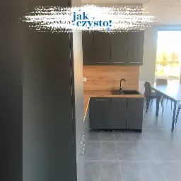 Wnętrze biurowe z nową, szarą zabudową kuchenną, drewnianym blatem i płytkami podłogowymi w odcieniach szarości. Za stołem widoczne krzesła.