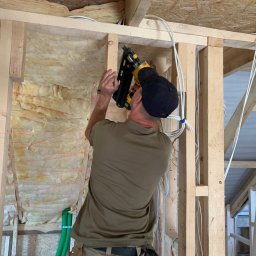 FIRMA USŁUGOWO-REMONTOWA - Pracownik w czapce mocuje drewniane elementy konstrukcji budynku za pomocą narzędzia pneumatycznego, widoczne kable elektryczne i izolacja z wełny mineralnej.