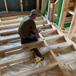 FIRMA USŁUGOWO-REMONTOWA - Pracownik w Jabłonce montuje drewnianą konstrukcję podłogi na poddaszu, widoczne narzędzia i zielone rury wentylacyjne.