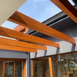 FIRMA USŁUGOWO-REMONTOWA - Drewniana pergola z belek w kolorze miodowym, zamontowana do ściany budynku w kolorze szarym, widok z dołu na niebieskie niebo, z oknami z żaluzjami w tle.