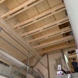 FIRMA USŁUGOWO-REMONTOWA - Wnętrze w trakcie prac wykończeniowych: drewniany strop z deskami i belkami, widoczne rusztowanie, drabina oparta o ścianę z białych pustaków, oraz pracownik w tle.