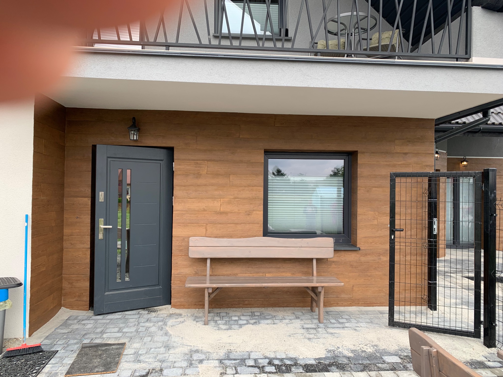 Elewacja budynku z drewnopodobnymi panelami, szara stolarka okienna i drzwiowa, ławka przed oknem oraz metalowe ogrodzenie.