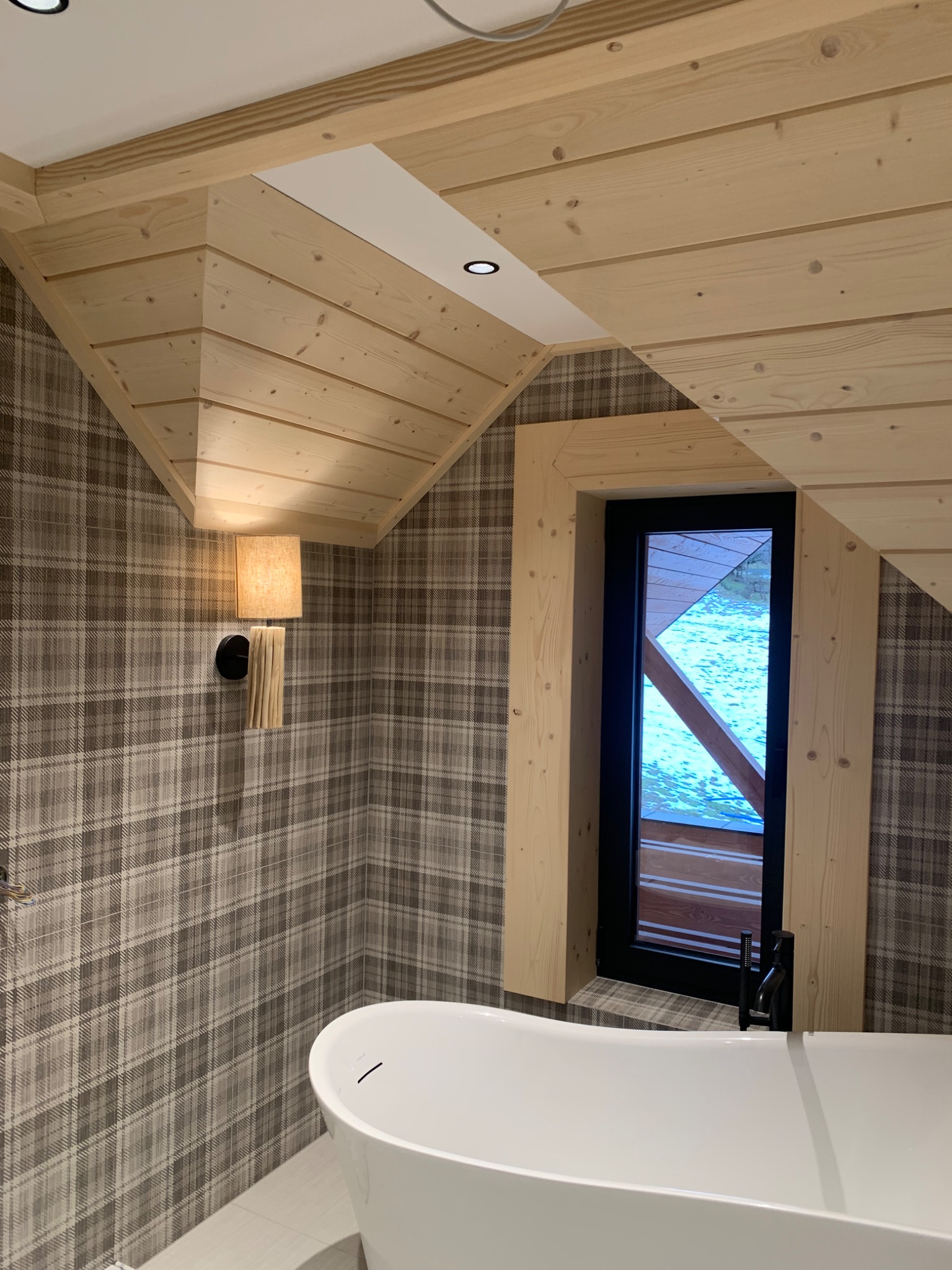 Nowoczesna łazienka w stylu góralskim z drewnianym sufitem, ścianą w kratę, wolnostojącą białą wanną i oknem z widokiem na zaśnieżony krajobraz.