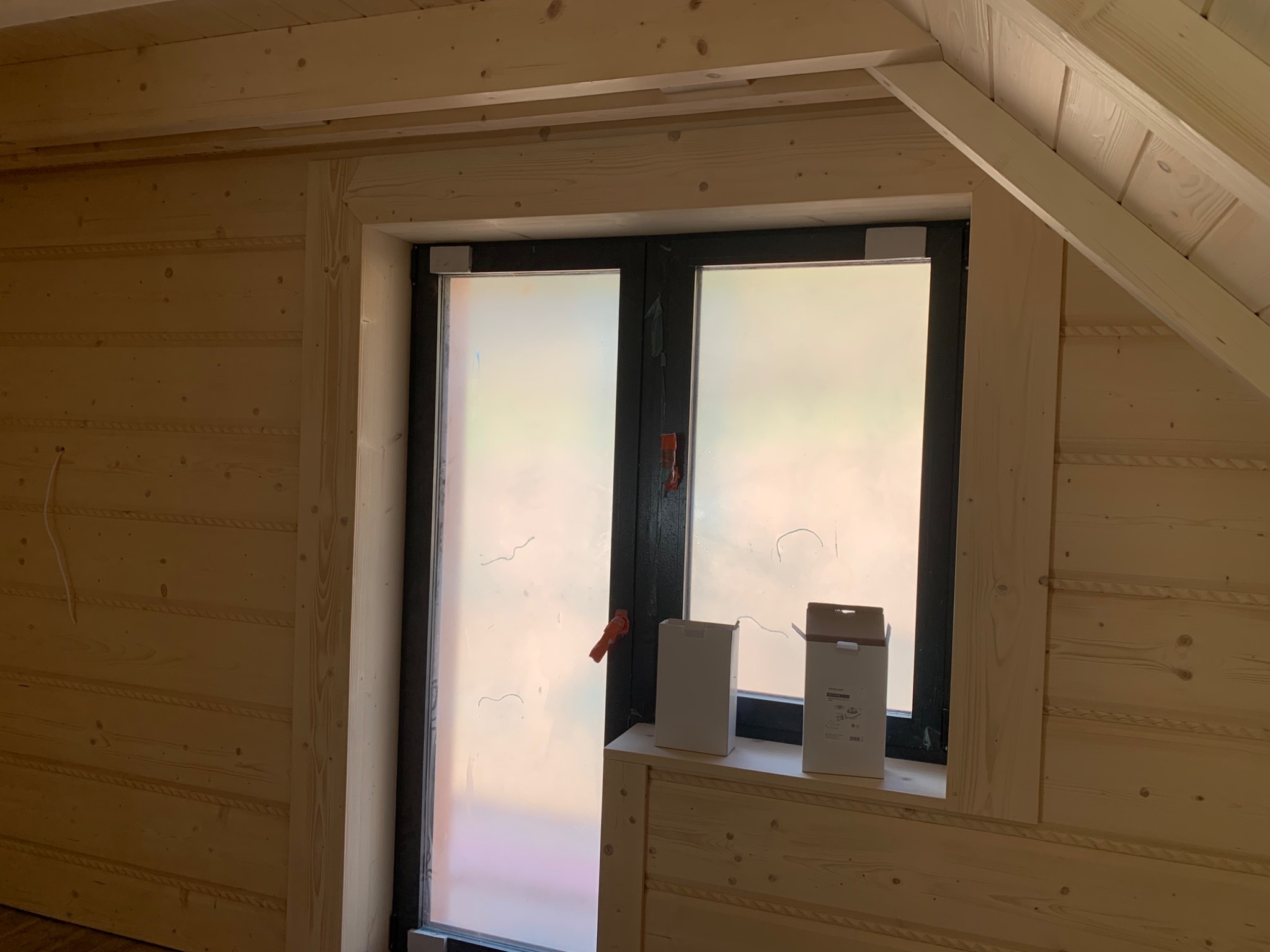 Wnętrze domu w trakcie wykańczania: drewniane panele ścienne z dekoracyjnymi listwami, okno z czarną ramą i matową szybą, parapet z dwiema paczkami materiałów budowlanych, widoczne elementy...