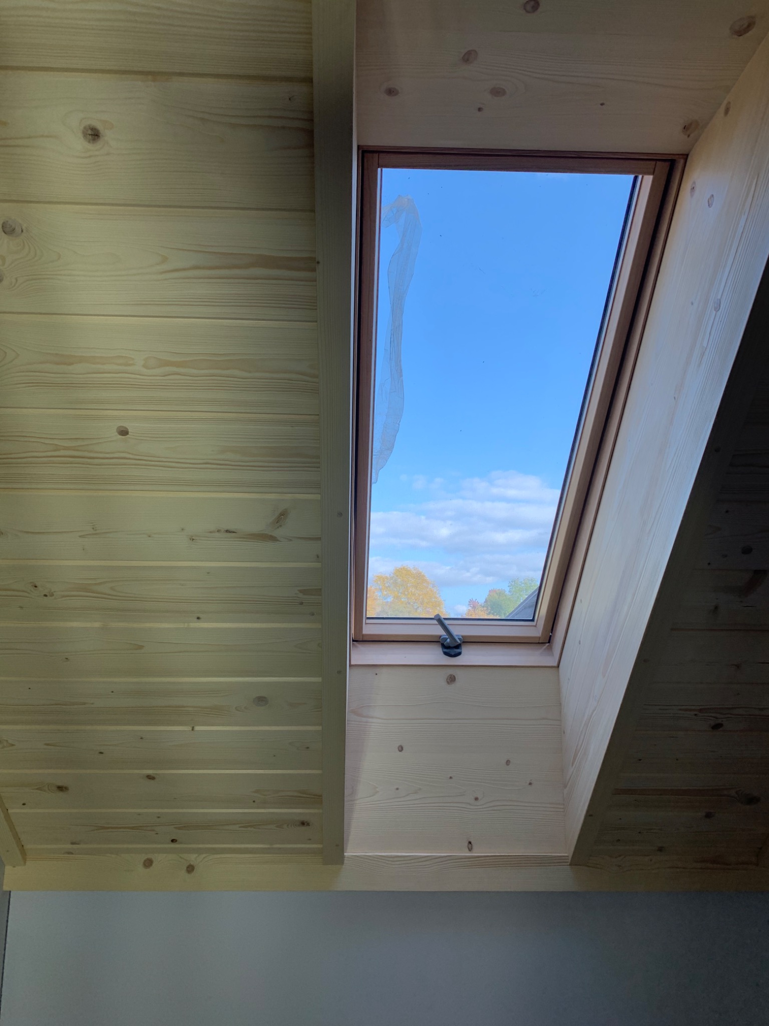 Drewniana boazeria i okno dachowe z widokiem na błękitne niebo w Jabłonce, z pozostałościami folii ochronnej na szybie.