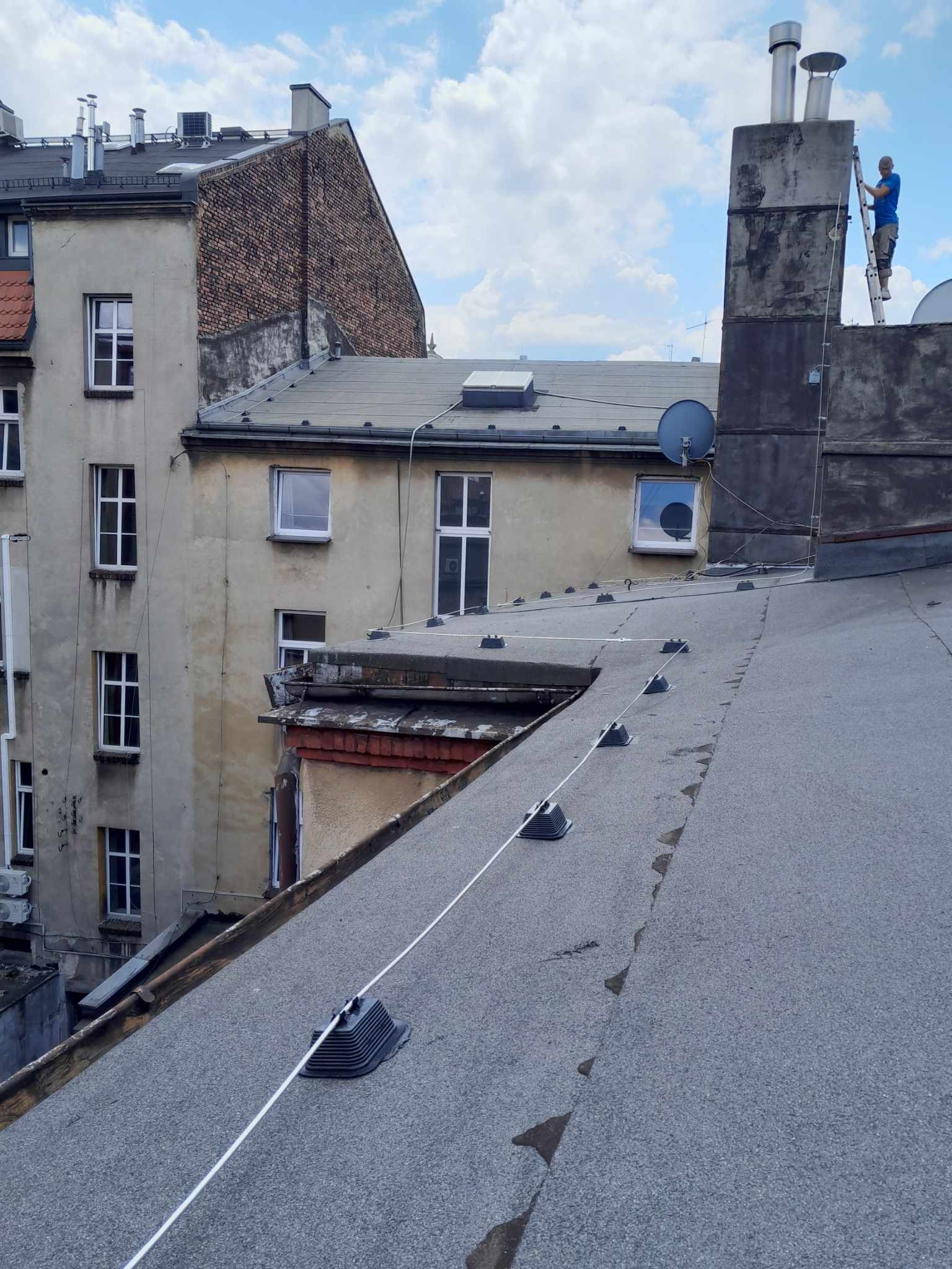 Instalacja odgromowa na dachu kamienicy, widoczny pracownik na drabinie przy kominie, przewody i uchwyty odgromowe na tle zabudowy miejskiej.