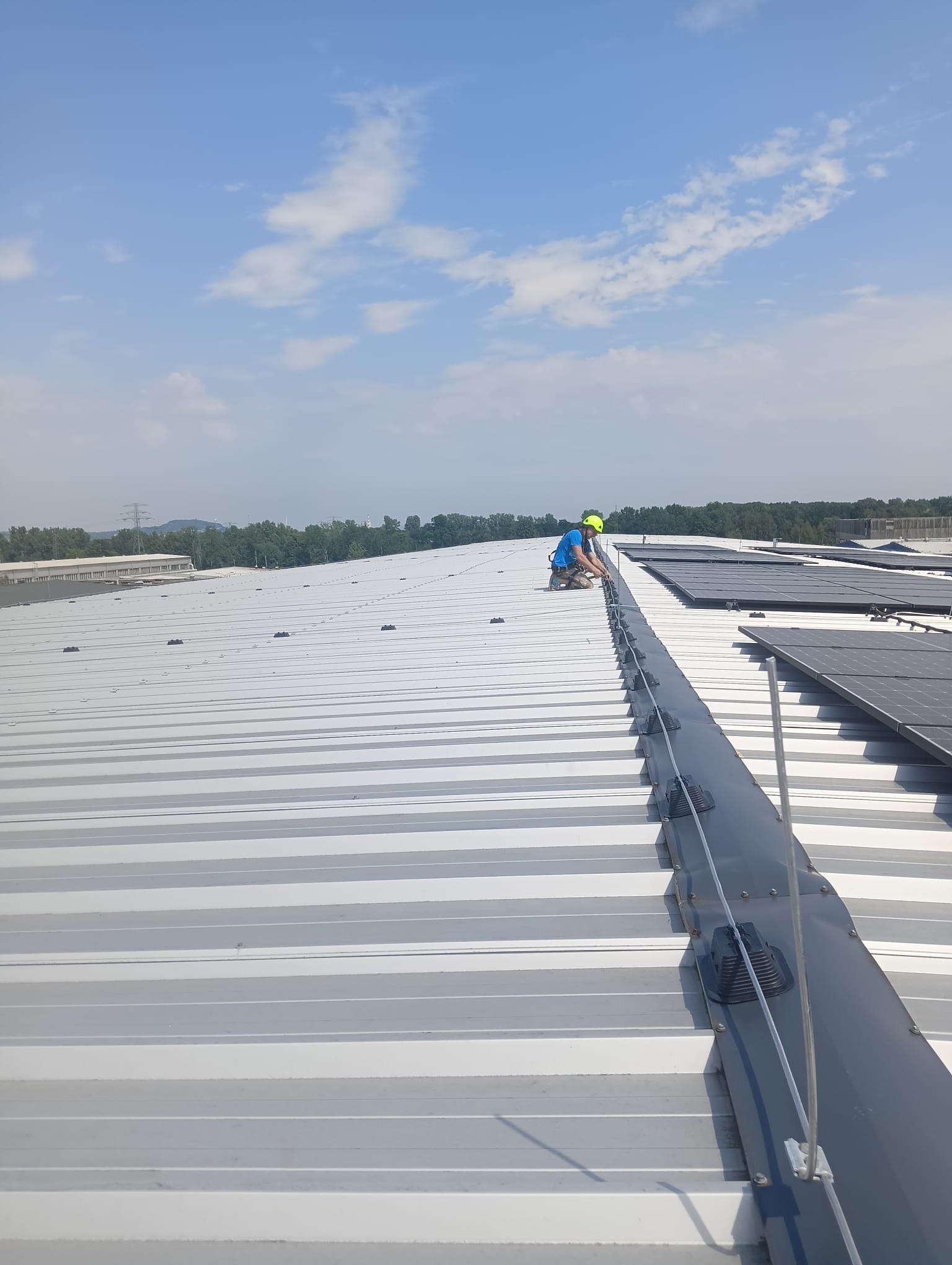 Pracownik w kasku i niebieskiej koszulce instaluje system odgromowy na dachu hali przemysłowej, widoczne panele słoneczne i uchwyty kabla odgromowego.
