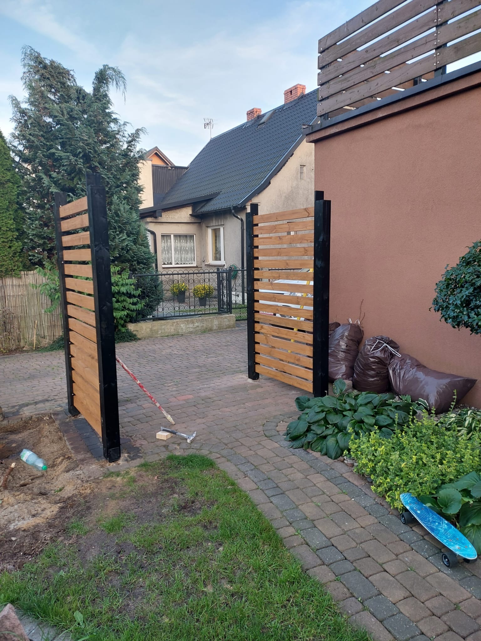 Montaż drewnianej bramy z poziomych desek z czarną ramą, widoczne narzędzia i worki z odpadami budowlanymi w ogrodzie.