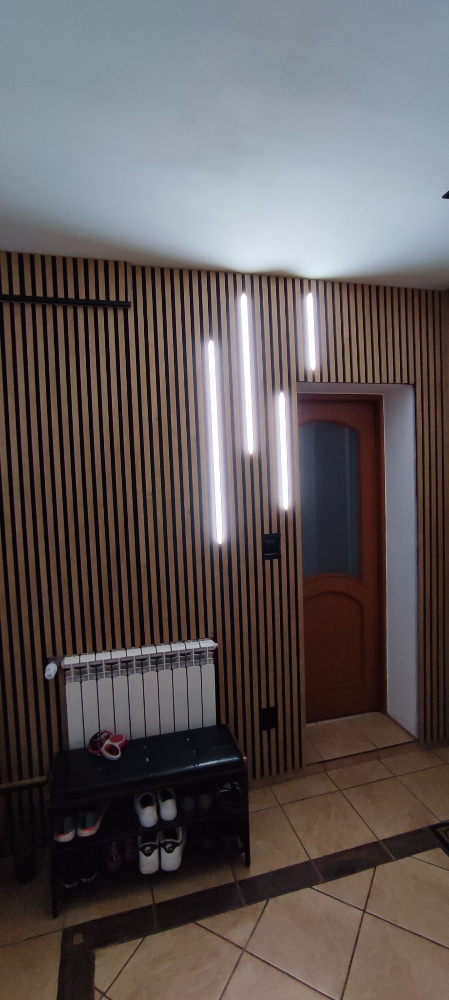 Nowoczesne wnętrze w Wróblowicach z drewnianą ścianą z pionowych paneli, oświetleniem LED, grzejnikiem i szafką na buty przy wejściu.