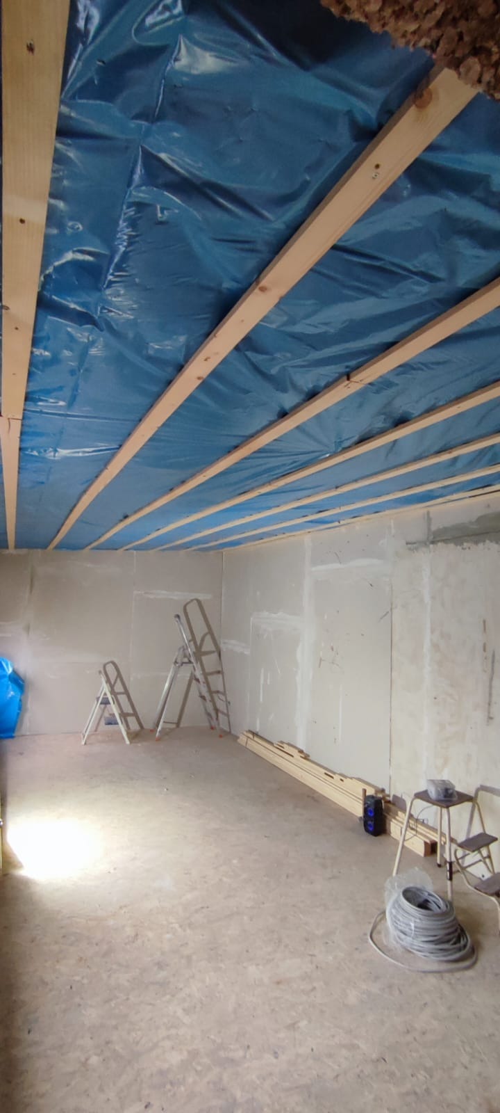 Wnętrze w trakcie prac wykończeniowych: sufit z izolacją zabezpieczoną folią, ściany z płyt gipsowo-kartonowych, drabiny i materiały budowlane na podłodze.