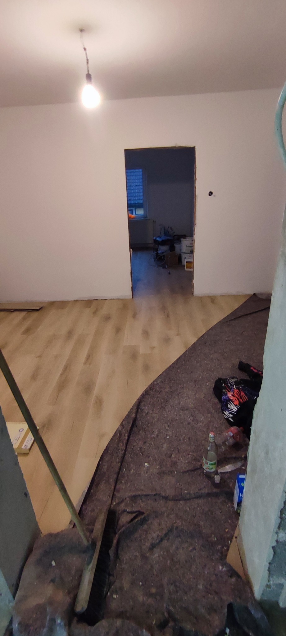 Wnętrze remontowanego pokoju z nową podłogą laminowaną, zabezpieczoną częściowo szarą folią ochronną; widoczna goła żarówka i otwór drzwiowy do kolejnego pomieszczenia z narzędziami i sprzętem.
