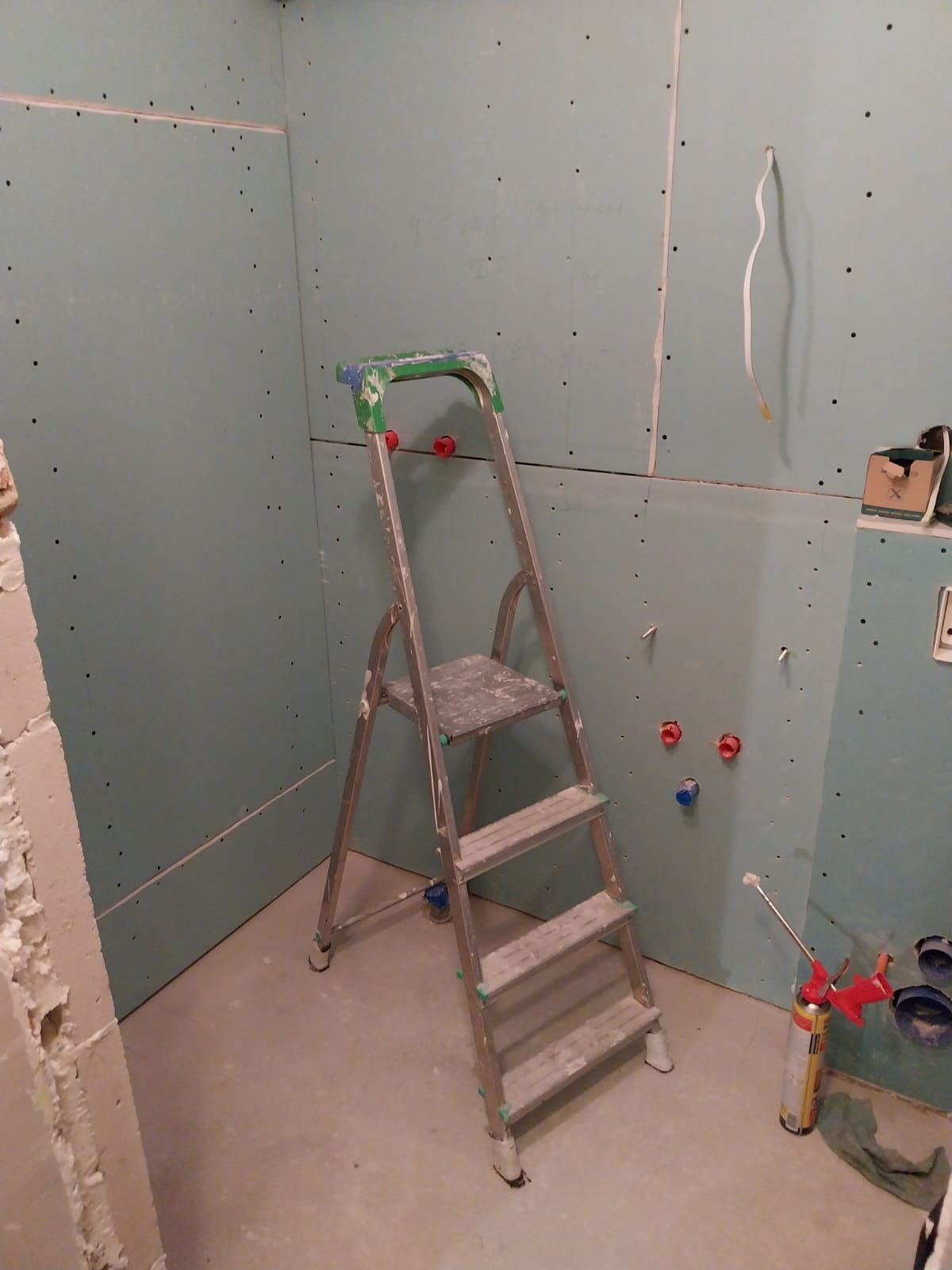 Wnętrze łazienki w trakcie remontu: ściany pokryte płytami gipsowo-kartonowymi, widoczna drabina aluminiowa z zielonymi elementami, wystające rury instalacji wodnej z czerwonymi i niebieskimi...