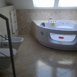 Jasna łazienka na poddaszu z beżowymi płytkami, białą wanną z hydromasażem, toaletą i bidetem, widoczne elementy metalowej balustrady schodów.