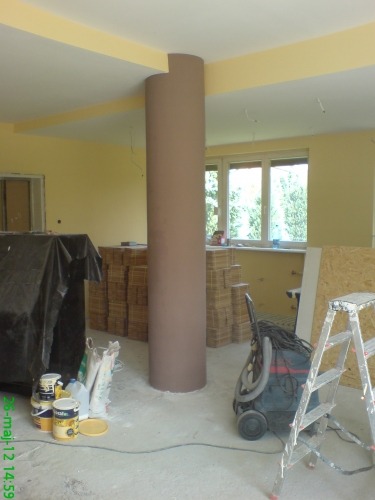 Wnętrze w trakcie prac wykończeniowych: kolumna w kolorze beżowym, ściany pomalowane na żółto, widoczne materiały budowlane, drabina i odkurzacz przemysłowy.