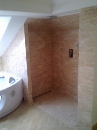 Ukończona kabina prysznicowa z beżowych płytek imitujących marmur, z odpływem liniowym i baterią podtynkową, obok narożna wanna z hydromasażem w łazience na poddaszu.