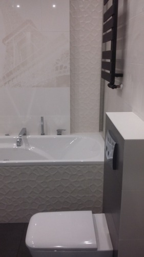 Nowoczesna łazienka z wanną obudowaną płytkami o trójwymiarowej fakturze, wiszącą toaletą i grzejnikiem drabinkowym w kolorze czarnym.