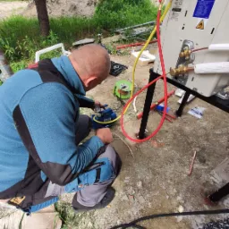 Instalator w niebieskiej bluzie podłącza pompę próżniową do zewnętrznej jednostki klimatyzacji na metalowym stojaku, widoczne miedziane rurki i przewody, tło z zielenią.