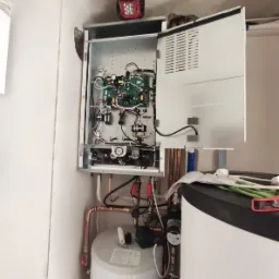 Otwarty panel kotła gazowego z widoczną elektroniką, pod nim zbiornik i miedziane rury, na górze lampa robocza na magnesie.