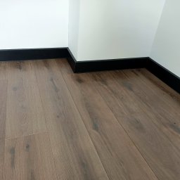 ParkietyWZ - Ułożona podłoga z paneli w odcieniach brązu z zamontowanymi czarnymi listwami przypodłogowymi, widoczny fragment białej ściany z otworem na gniazdko elektryczne.