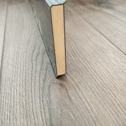 ParkietyWZ - Przekrój listwy przypodłogowej z MDF na tle paneli podłogowych z wyraźną strukturą drewna.