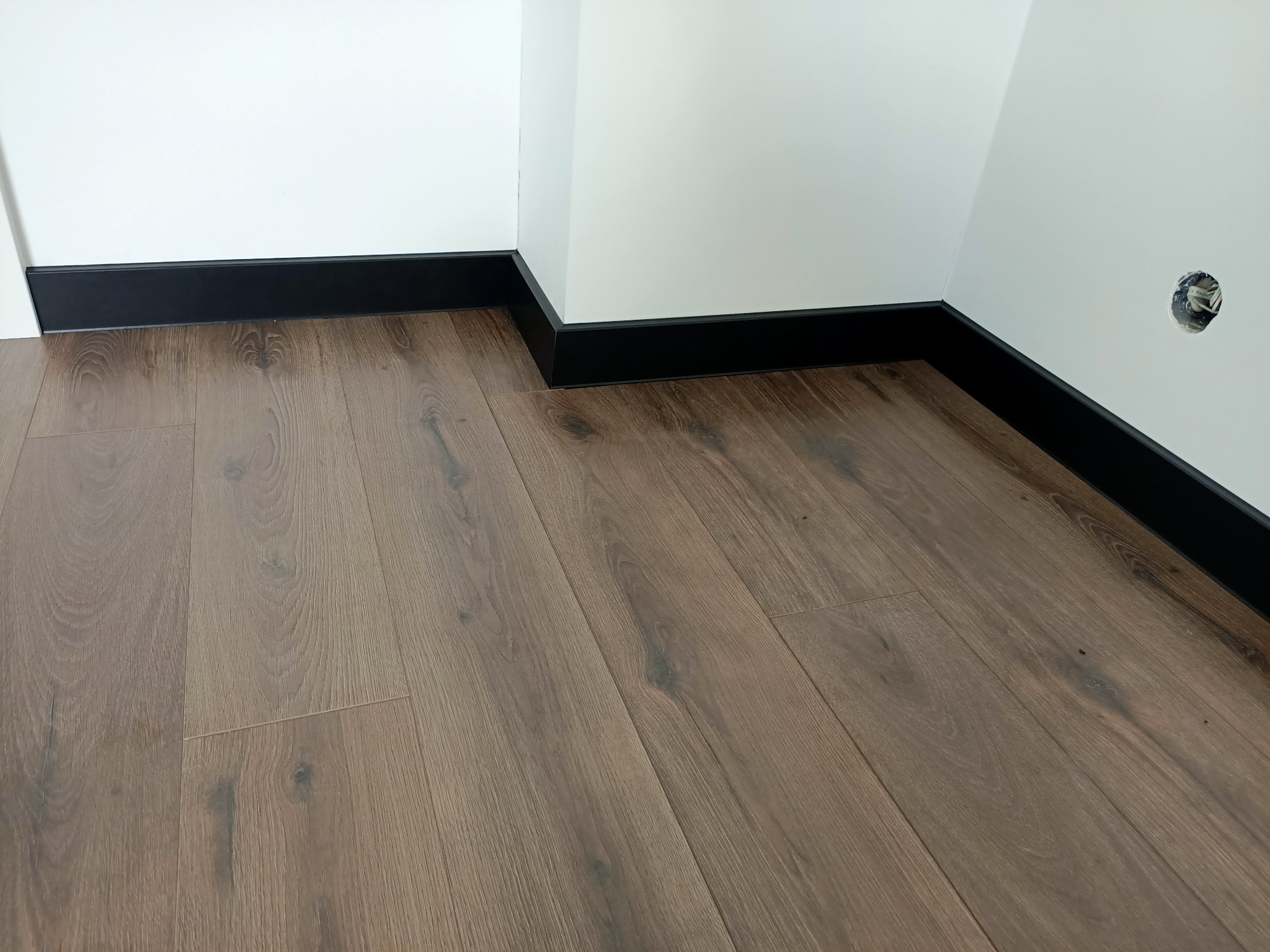 Ułożona podłoga z paneli w odcieniach brązu z zamontowanymi czarnymi listwami przypodłogowymi, widoczny fragment białej ściany z otworem na gniazdko elektryczne.