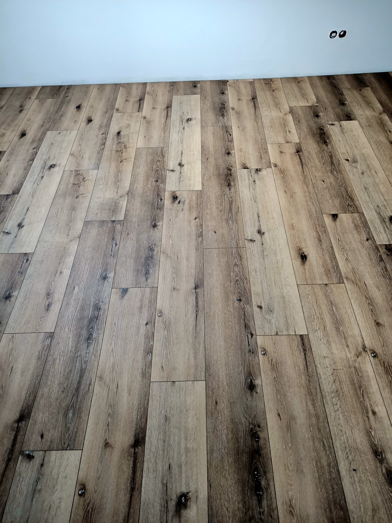 Ułożona podłoga z paneli imitujących postarzane drewno w odcieniach brązu i beżu, widoczne gniazdka elektryczne na białej ścianie.