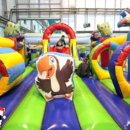 Dzieci bawią się na kolorowym, dmuchanym placu zabaw z motywami zwierzęcymi, w hali z metalową konstrukcją sufitu.