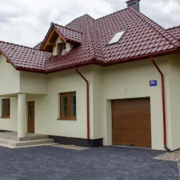 Nowoczesny dom jednorodzinny z czerwoną dachówką, beżową elewacją i drewnianą bramą garażową, widok z zewnątrz na kostkę brukową i numer domu 58a.