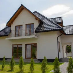 Biały dom z ciemną dachówką, balkonem i dużymi oknami, otoczony trawnikiem z młodymi tujami.