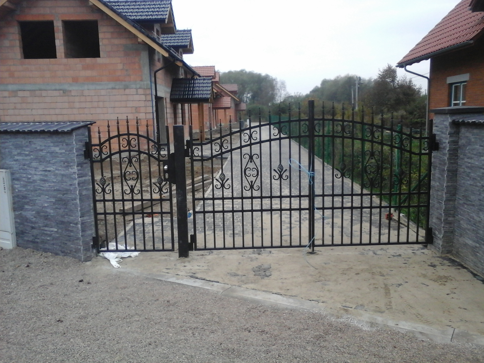 Czarna, metalowa brama wjazdowa z dekoracyjnymi elementami, osadzona między kamiennymi słupkami, prowadząca do posesji z domem w stanie surowym otwartym.