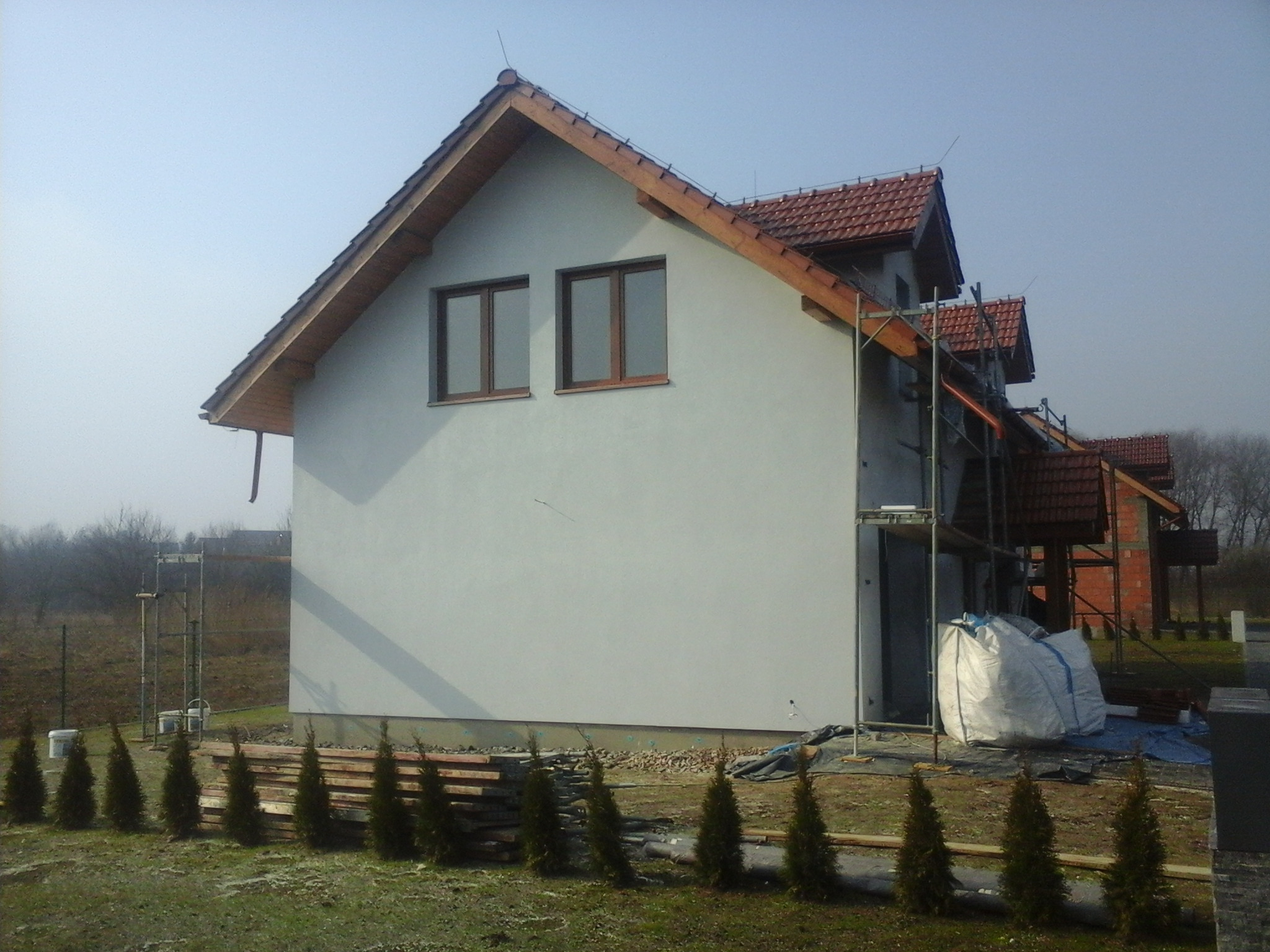Surowy stan elewacji domu jednorodzinnego z dwuspadowym dachem pokrytym czerwoną dachówką, widoczne rusztowania, worki z materiałami budowlanymi i rząd małych tujek posadzonych wzdłuż ściany.