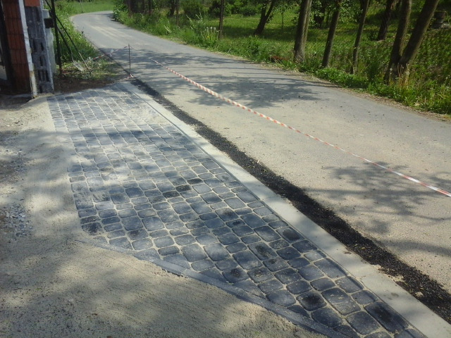 Układanie kostki brukowej w odcieniach szarości, widoczne obrzeża i taśma ostrzegawcza, teren w trakcie prac ziemnych, droga asfaltowa w tle.