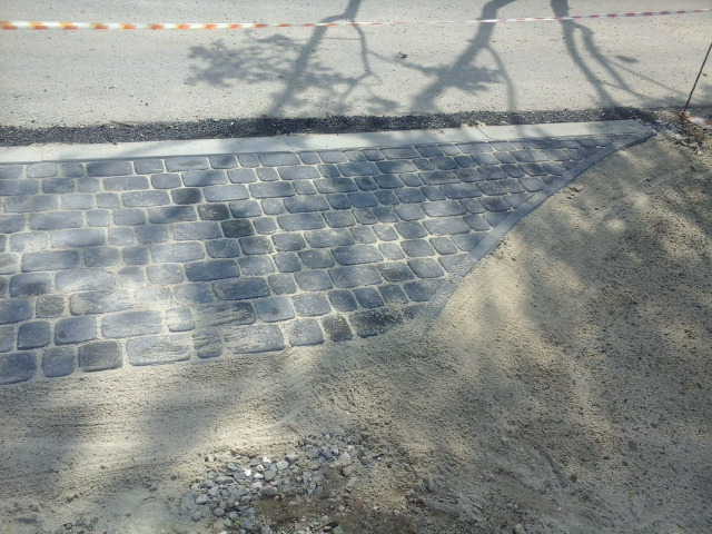 Ułożona kostka brukowa w szarym odcieniu, widoczne piaskowe podsypanie i cień drzewa na nawierzchni.