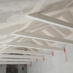Mariusz Chmielewski Usługi Remontowo-Wykończeniowe - Skośny sufit z płyt kartonowo-gipsowych w trakcie szpachlowania, widoczne białe drewniane belki konstrukcyjne i pomarańczowe elementy mocujące, ślady szpachli na ścianach.