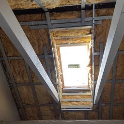 Mariusz Chmielewski Usługi Remontowo-Wykończeniowe - Montaż izolacji z wełny mineralnej na poddaszu, widoczne profile stalowe i drewniane belki, okno dachowe w tle.