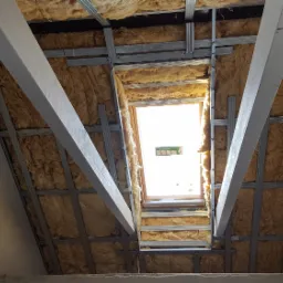 Montaż izolacji z wełny mineralnej na poddaszu, widoczne profile stalowe i drewniane belki, okno dachowe w tle.