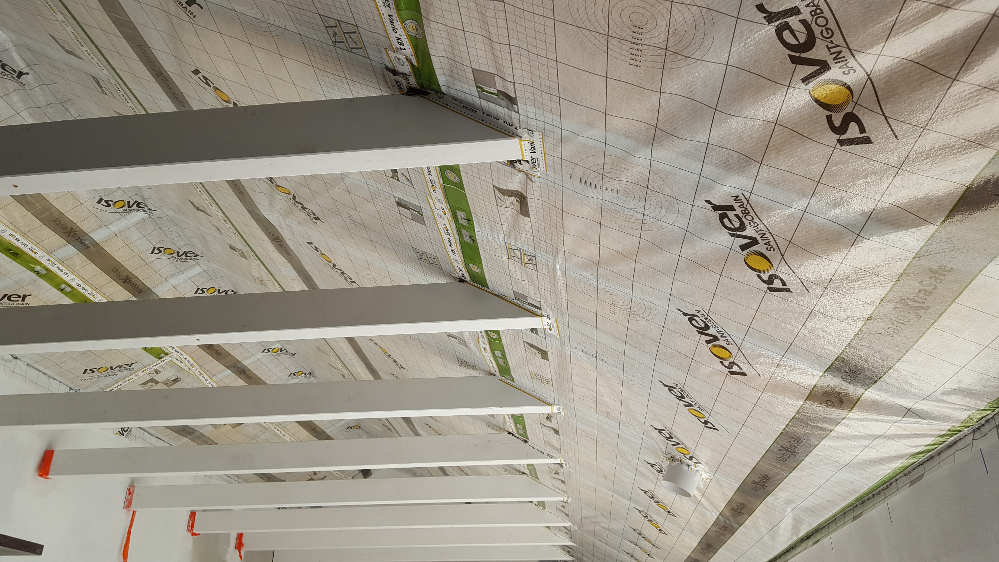 Montaż izolacji Isover Vario XtraSafe na poddaszu, widoczne białe belki konstrukcyjne i folia paroizolacyjna z nadrukowanym logo Isover.