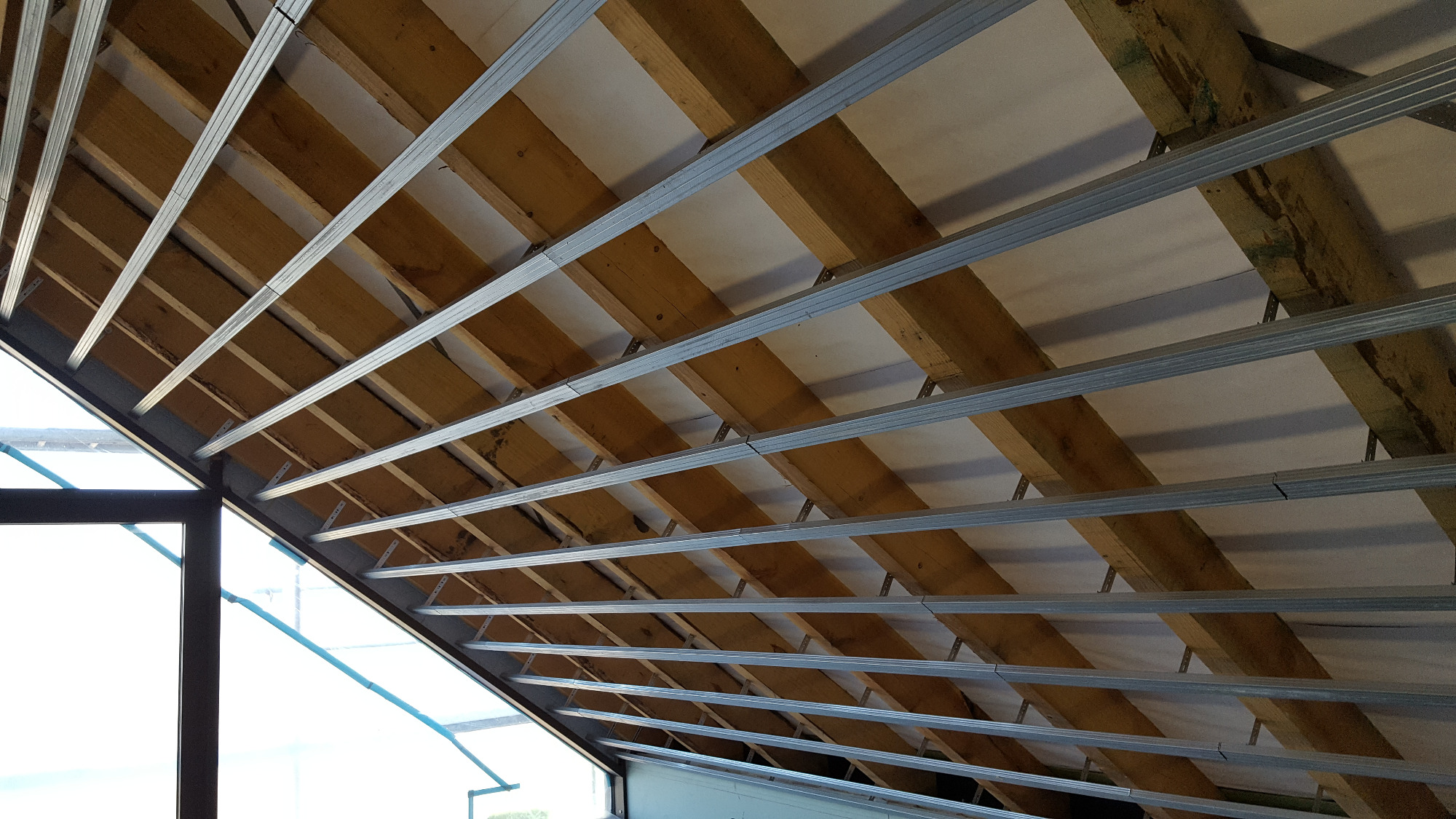 Konstrukcja sufitu podwieszanego z profili stalowych i drewnianych belek, widok od dołu, oświetlona naturalnym światłem wpadającym przez okno.