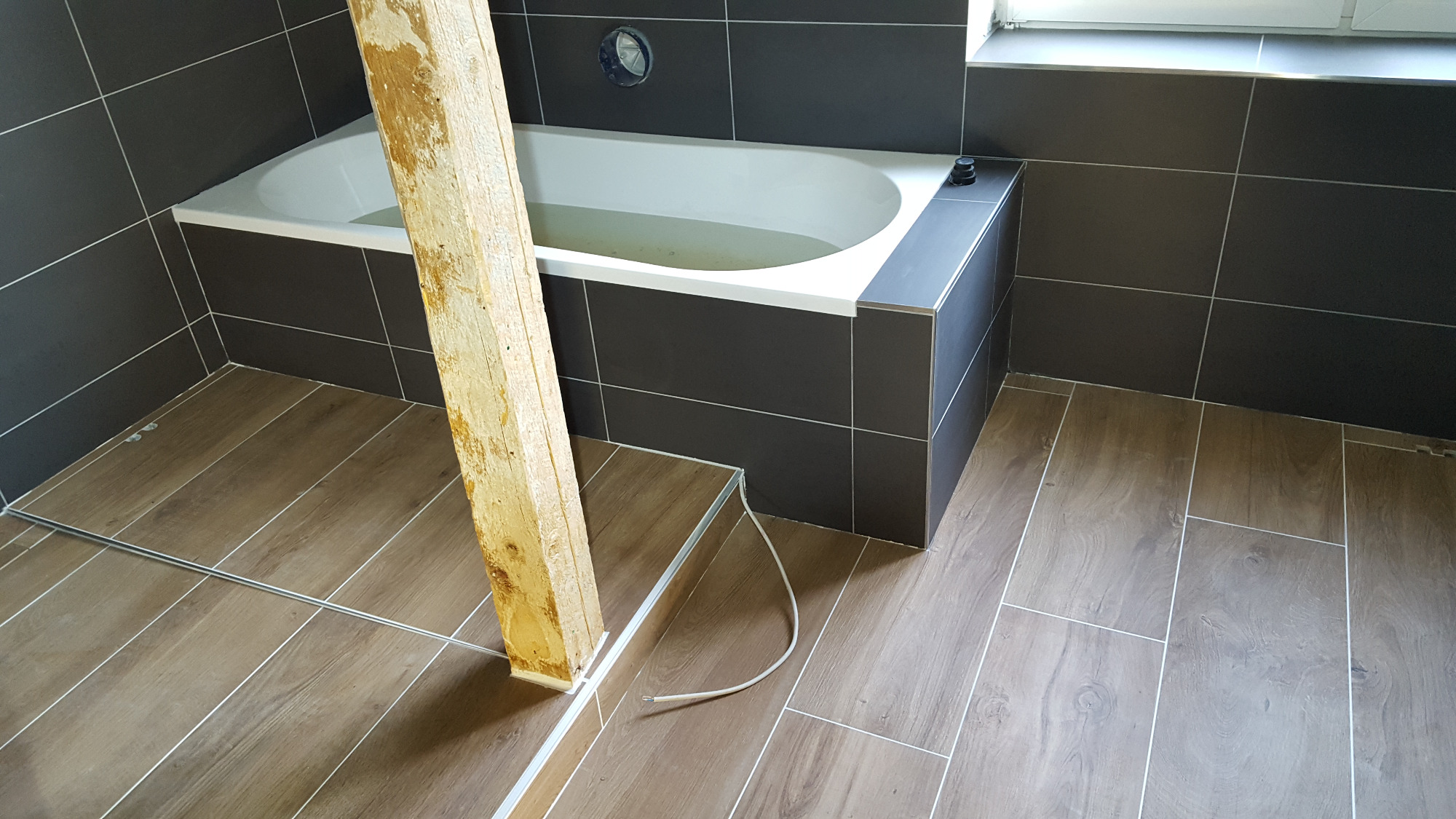 Wnętrze łazienki w trakcie remontu: widoczna wanna z szarą obudową i ciemnymi płytkami na ścianach, drewniana belka podpierająca oraz podłoga wyłożona panelami imitującymi drewno.